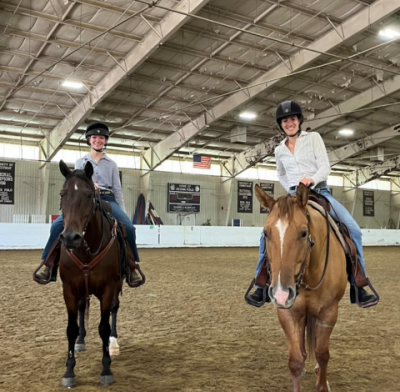 Two students posing for photo while riding horses in arena.